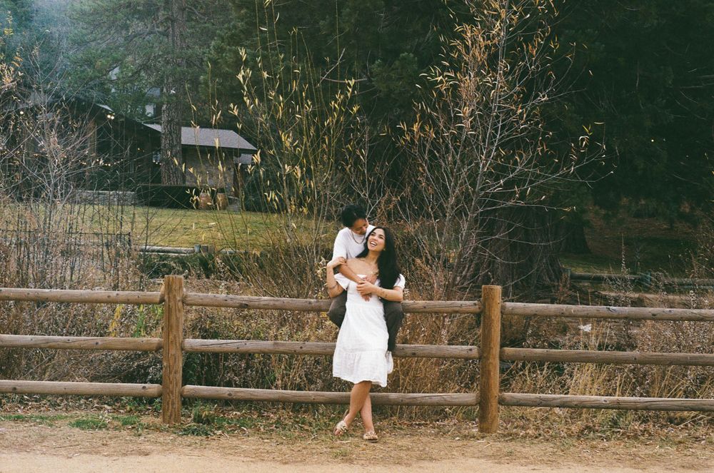A couple embraces by a rustic wooden fence in a countryside setting on a vintage-style film photograph.