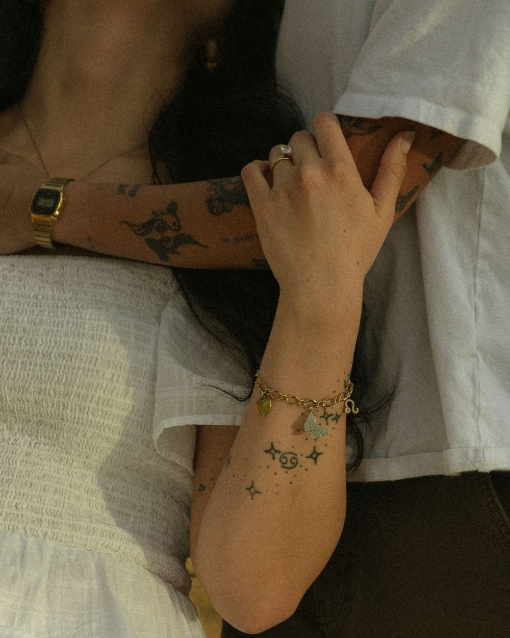 Two arms with delicate butterfly tattoos and jewelry reach towards each other against a light fabric background.
