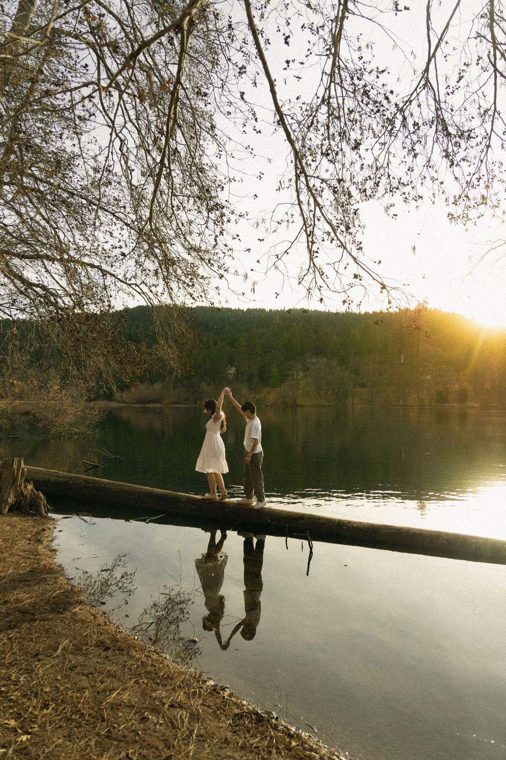 Two people stand on a fallen log over a serene lake at sunset, their reflection mirrored in the calm water.