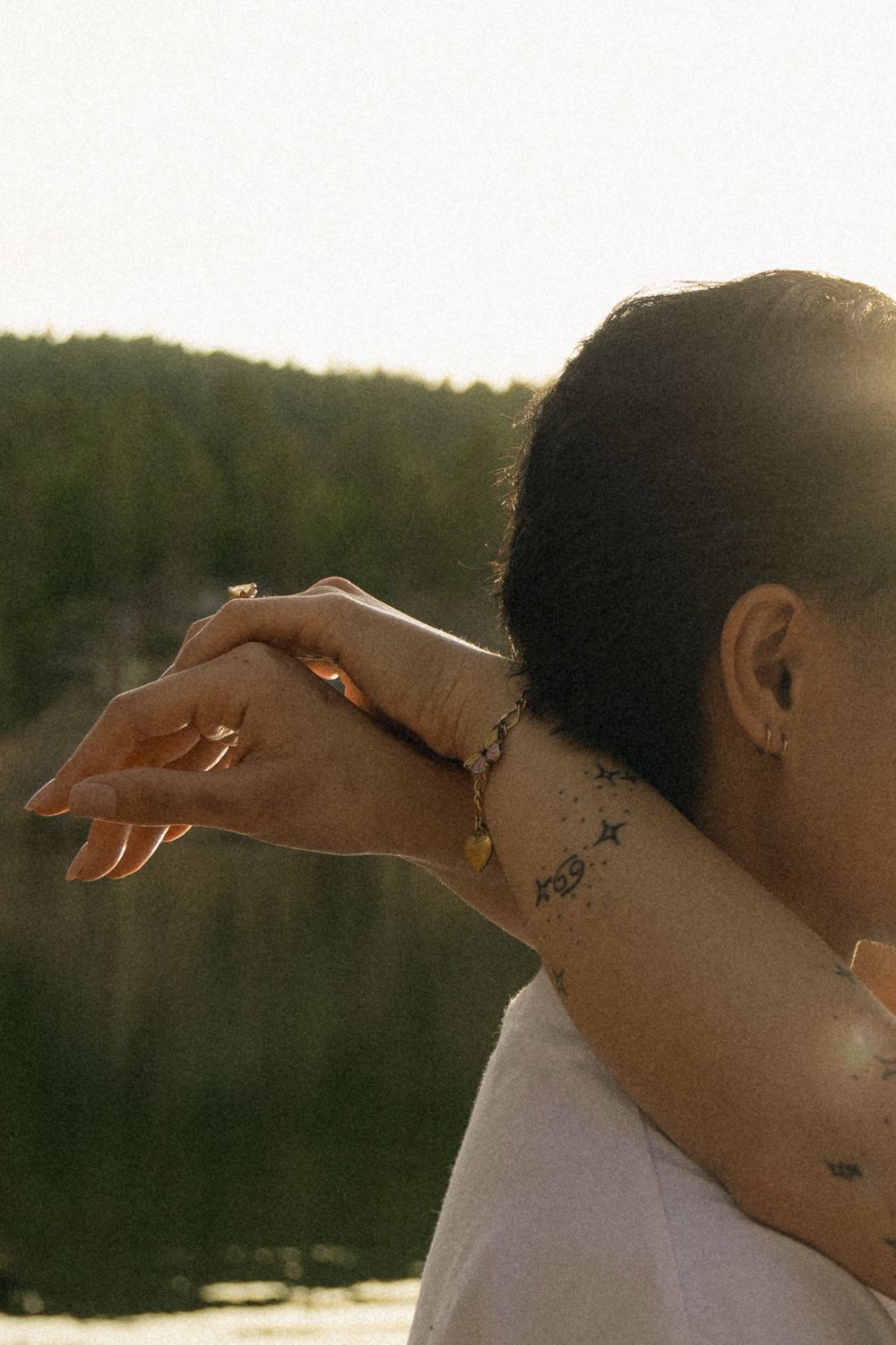 Close-up view of a person with tattoos showing a delicate hand gesture against a sunlit forest backdrop.