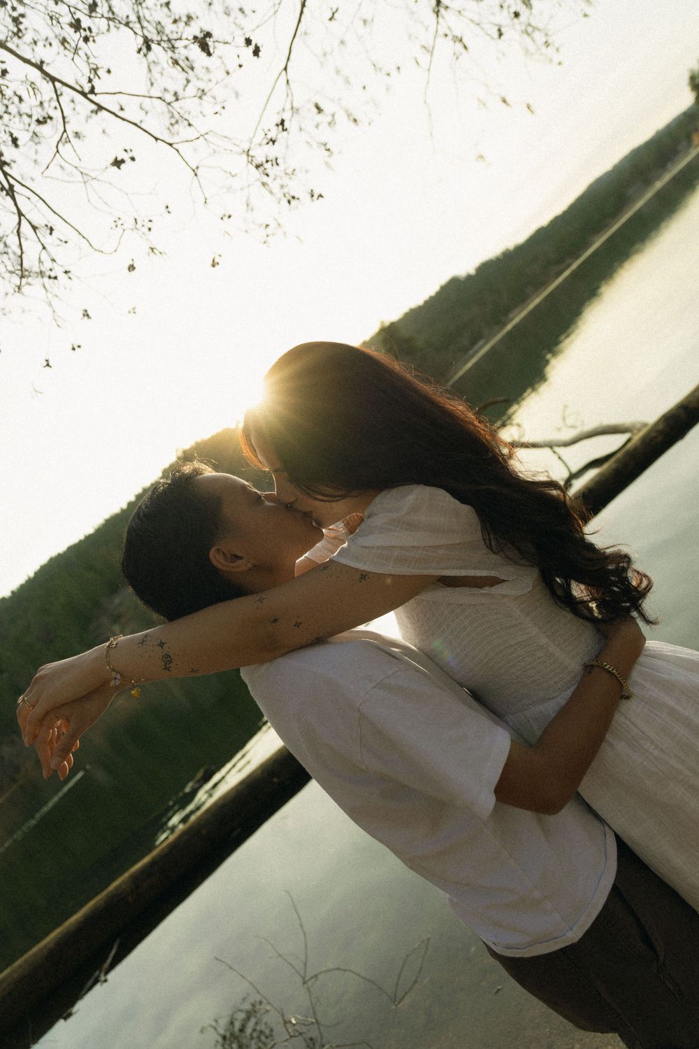 A romantic couple embraces against a sunset backdrop near water, both dressed in white clothing.