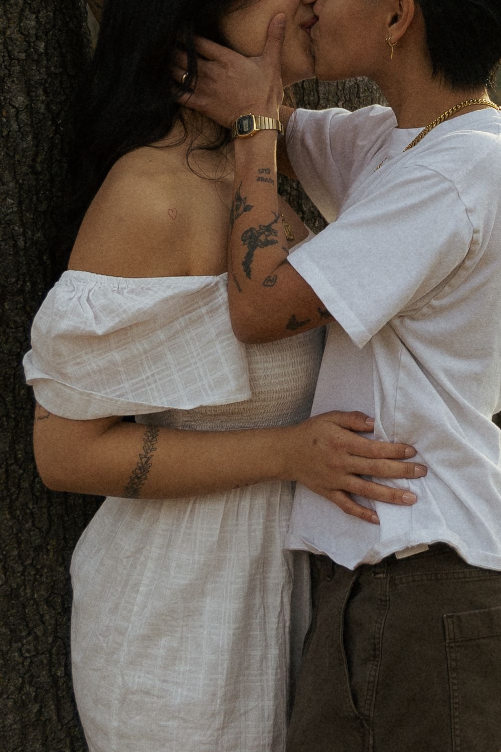 Two people in white clothing embrace intimately against a tree trunk showing visible tattoos.