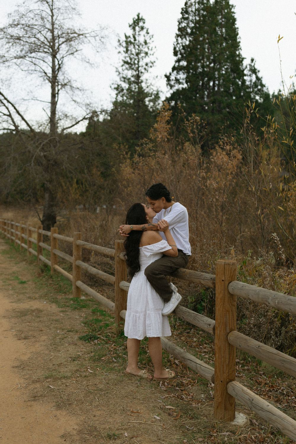 A couple sharing an intimate moment while sitting on a wooden fence along a dirt path surrounded by autumn foliage.