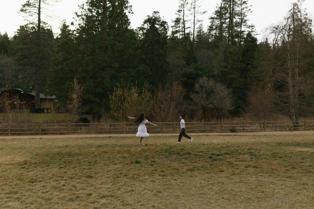 Two people in white clothing walk across a brown grassy field with evergreen trees in the background.
