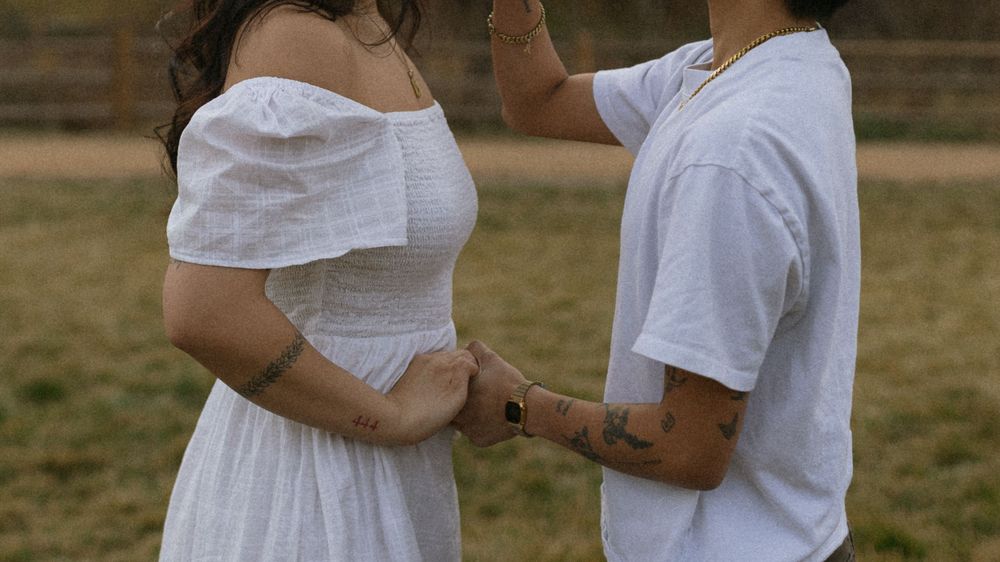 A couple in white outfits holding hands in an outdoor field, sharing an intimate moment together.