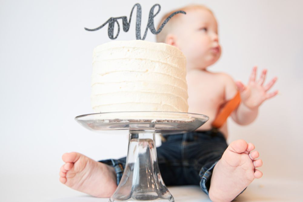 White cake with 'one' text topper on crystal stand against neutral background.
