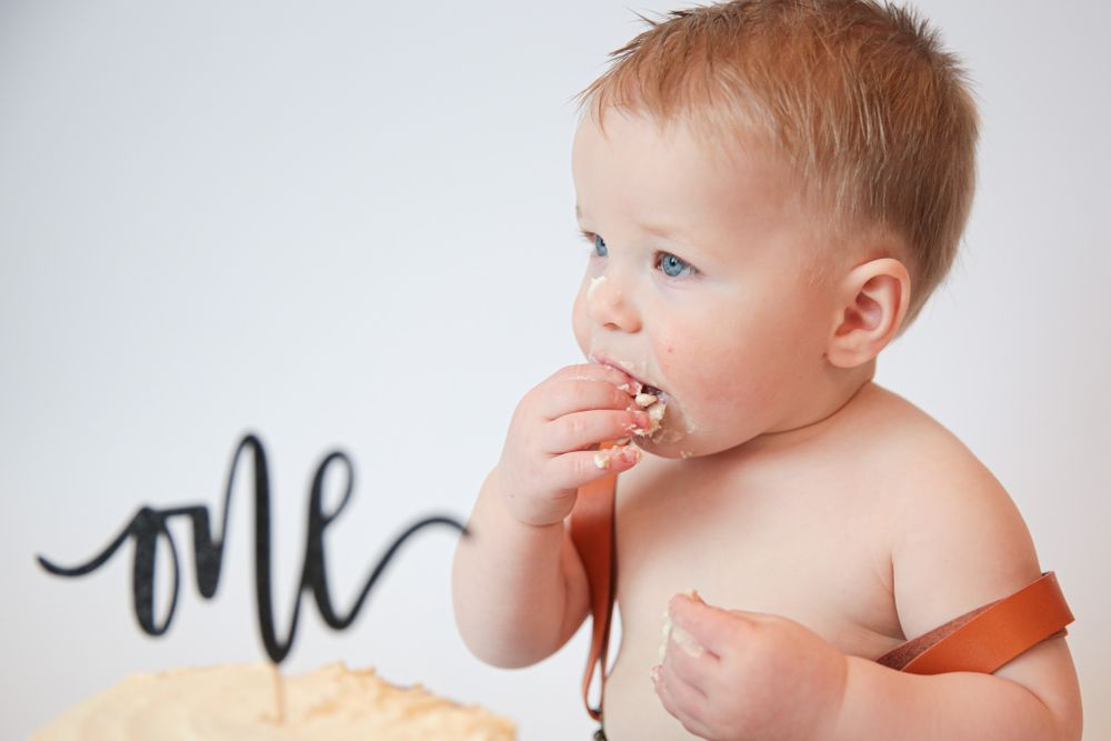 Small hands reach for cake crumbs next to black 'one' decoration on white background.