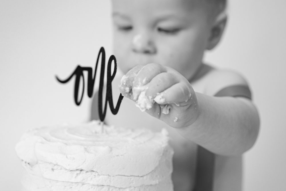 Black and white photo of tiny hands touching white frosted cake with 'one' decoration.
