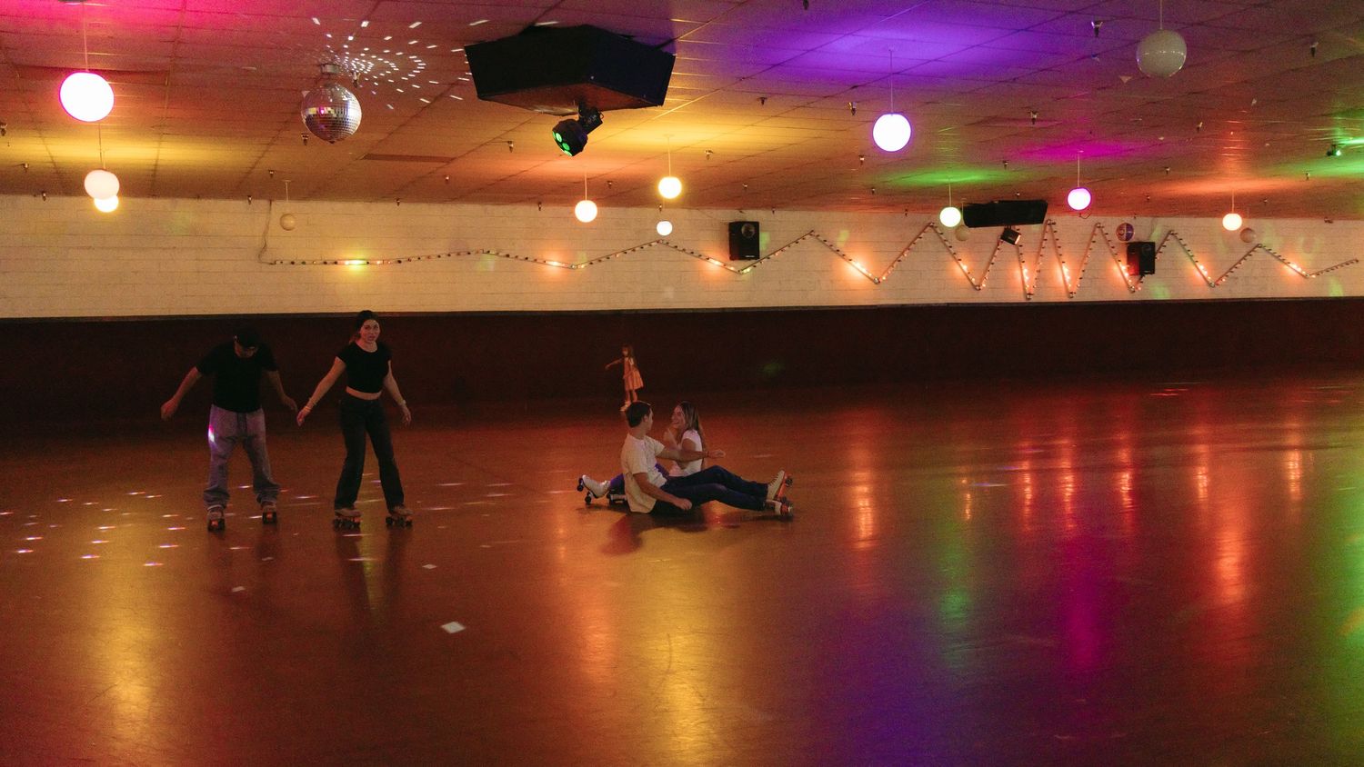 People skating at a roller rink with colorful lighting and reflective floor casting vibrant neon glows.