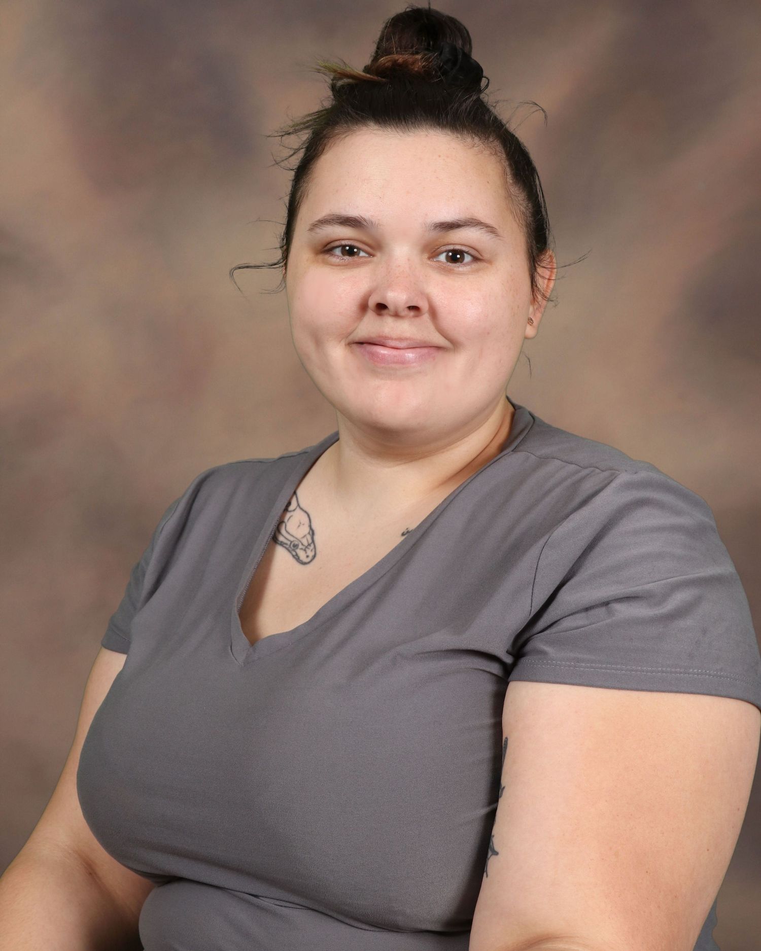 Professional studio portrait of a medical worker wearing grey scrubs with hair in a bun against a brown background.