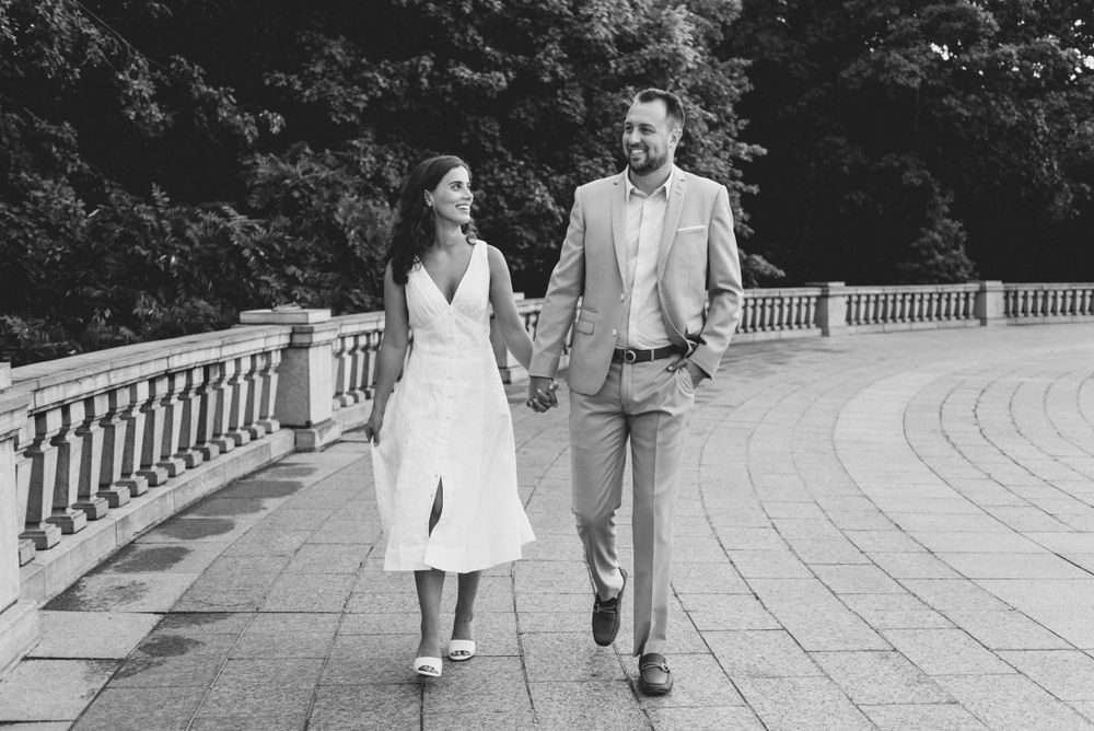 A couple strolls together along a curved stone balustrade walkway in an elegant black and white photograph.