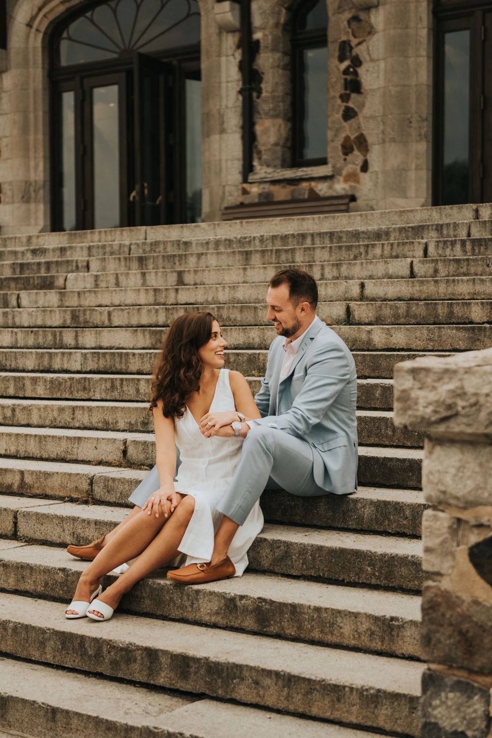 A romantic couple sits together on stone steps of a historic building in elegant formal attire.
