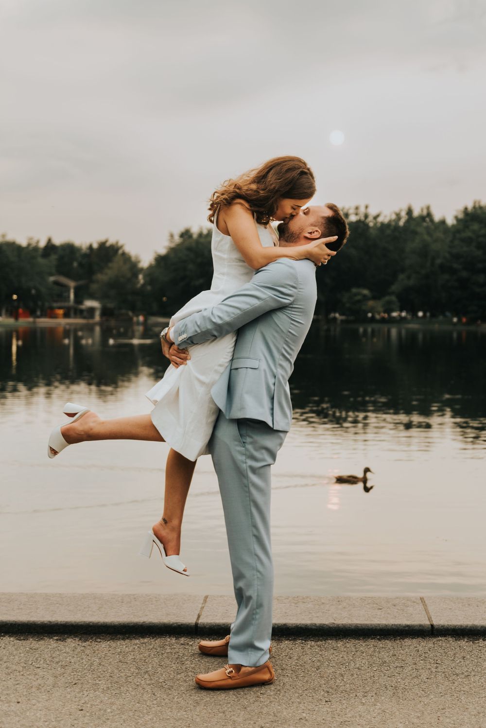 Romantic couple embraces by a serene lake at dusk with trees in background.