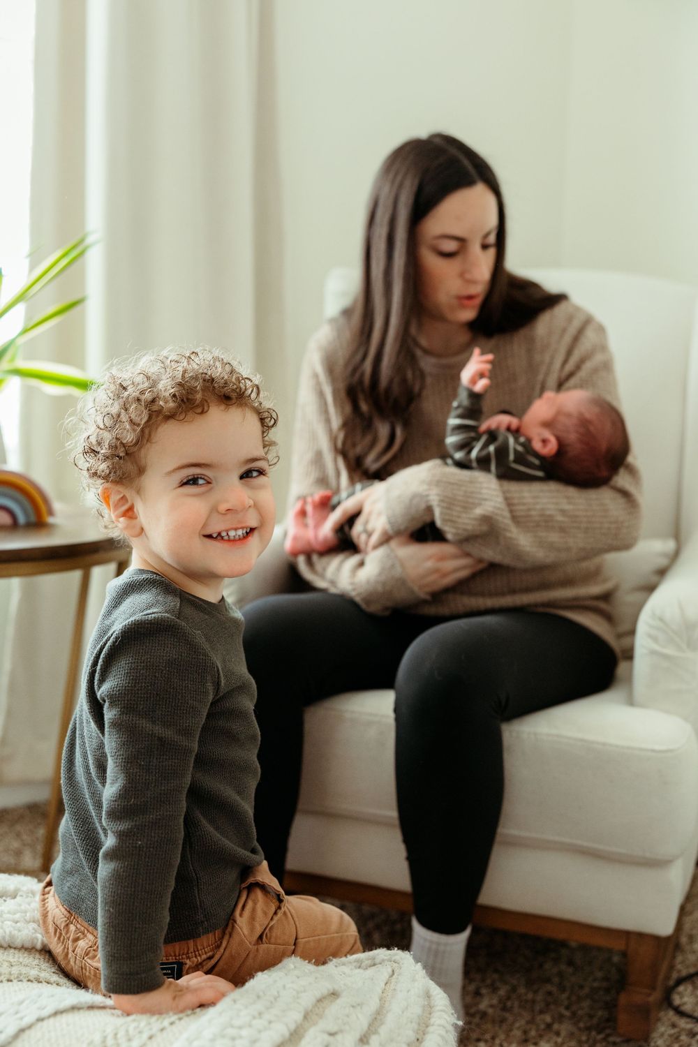 A young child with curly hair sits on a carpet while someone holds a baby on a couch in a bright living room.