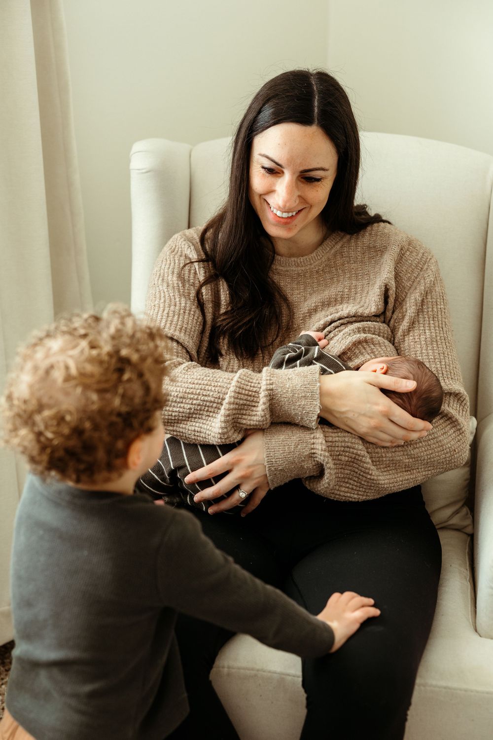 Woman in beige sweater sitting in white armchair sharing tender moment with curly-haired toddler.