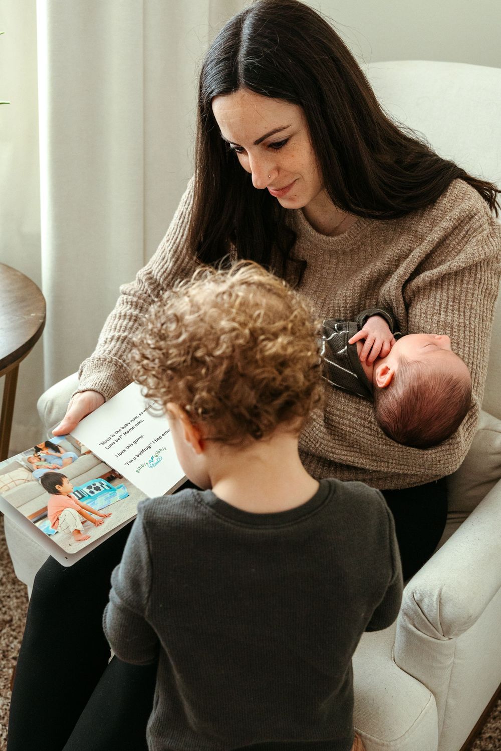 Family reading time in cozy armchair with toddler and infant while wearing comfortable knits.