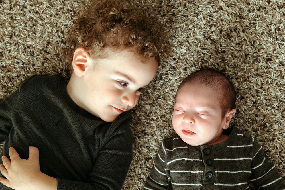 A young child and an infant lie together on a beige carpet, with the older child wearing a black sweater and the baby in stripes.