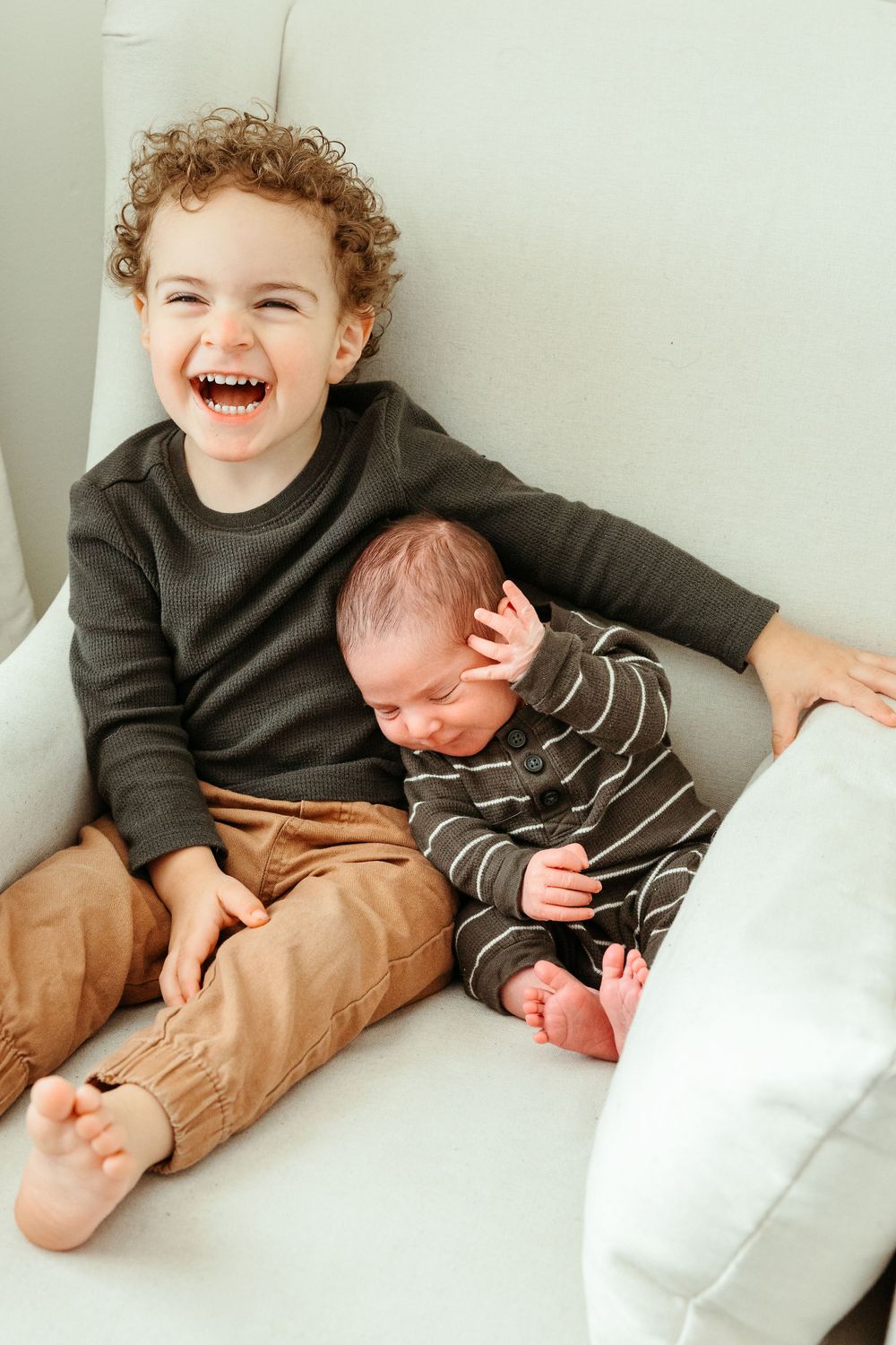 Two children sitting on white couch with curly-haired child laughing while younger sibling leans over.