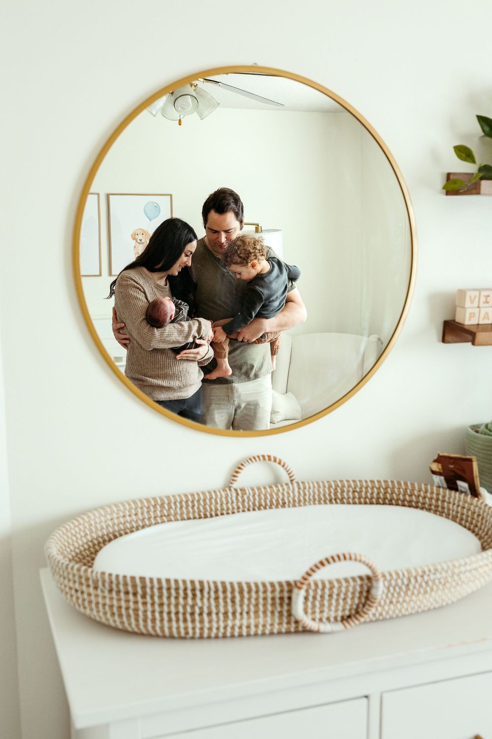 A round mirror on a white wall reflects a family moment, with a woven basket on the dresser below.