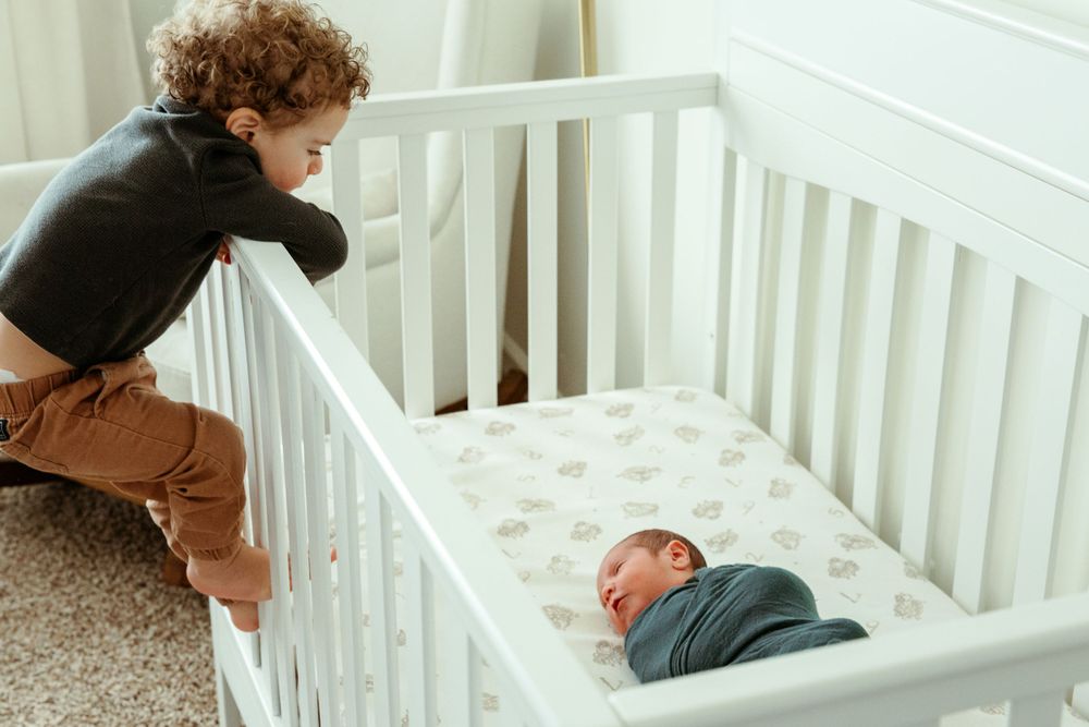 Young child curiously peers over white crib railing at baby lying in crib with patterned mattress.