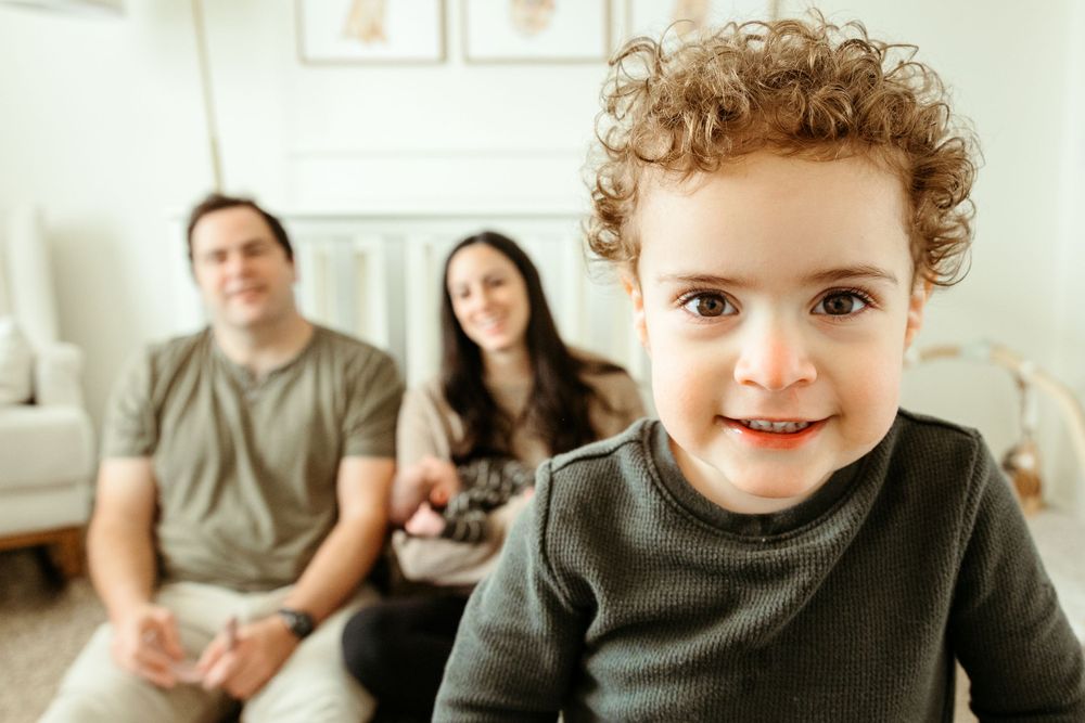 A young child with curly hair sits in the foreground while two adults relax on a couch in the background.
