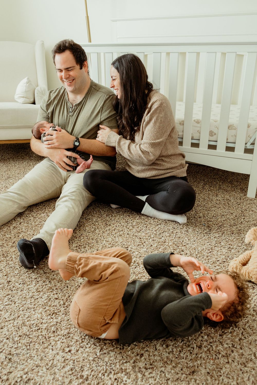 Family shares a fun moment together on a beige carpet in a nursery room with a white crib.