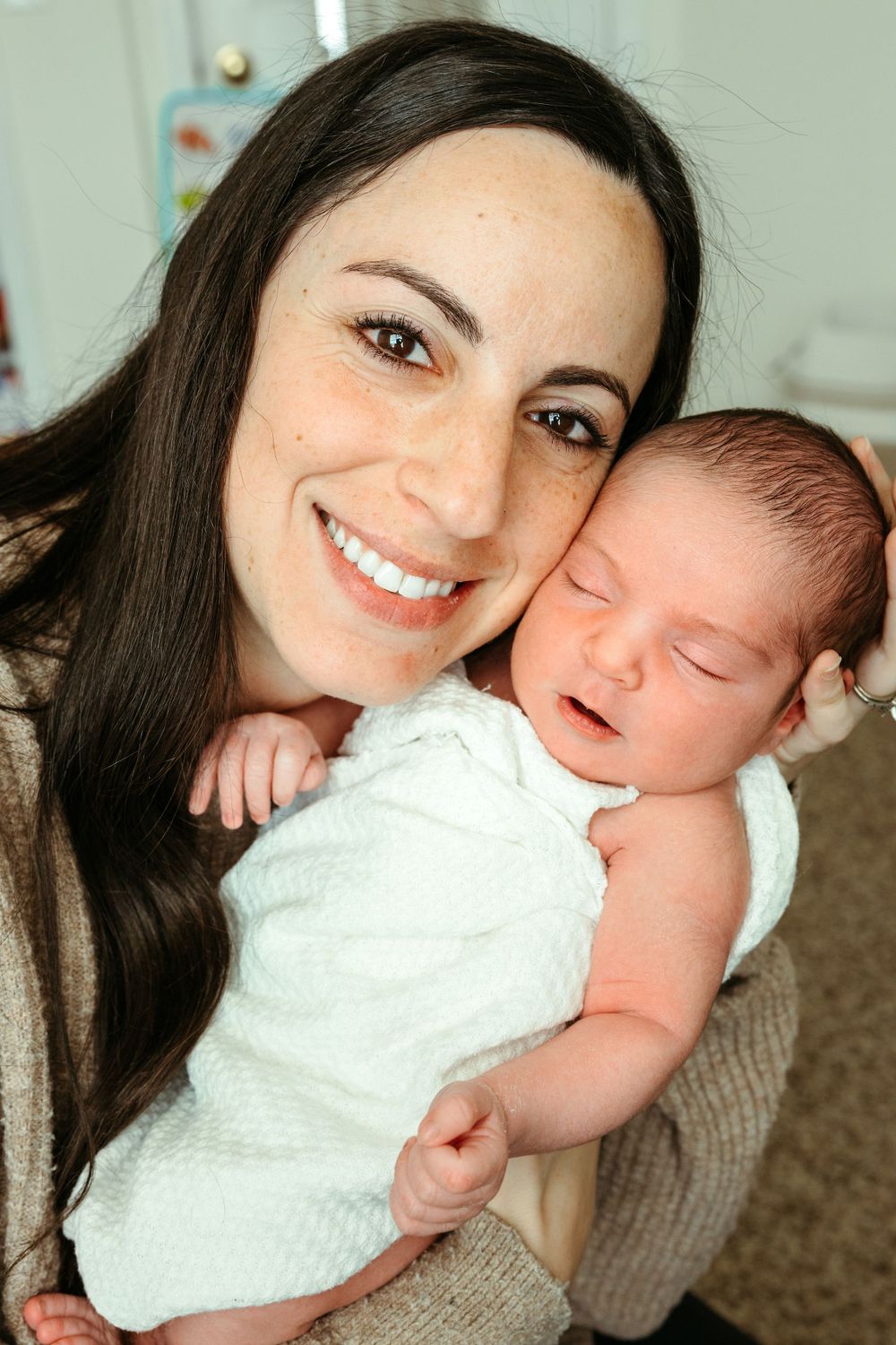 A woman with long dark hair holds a sleeping newborn baby dressed in white against her chest.