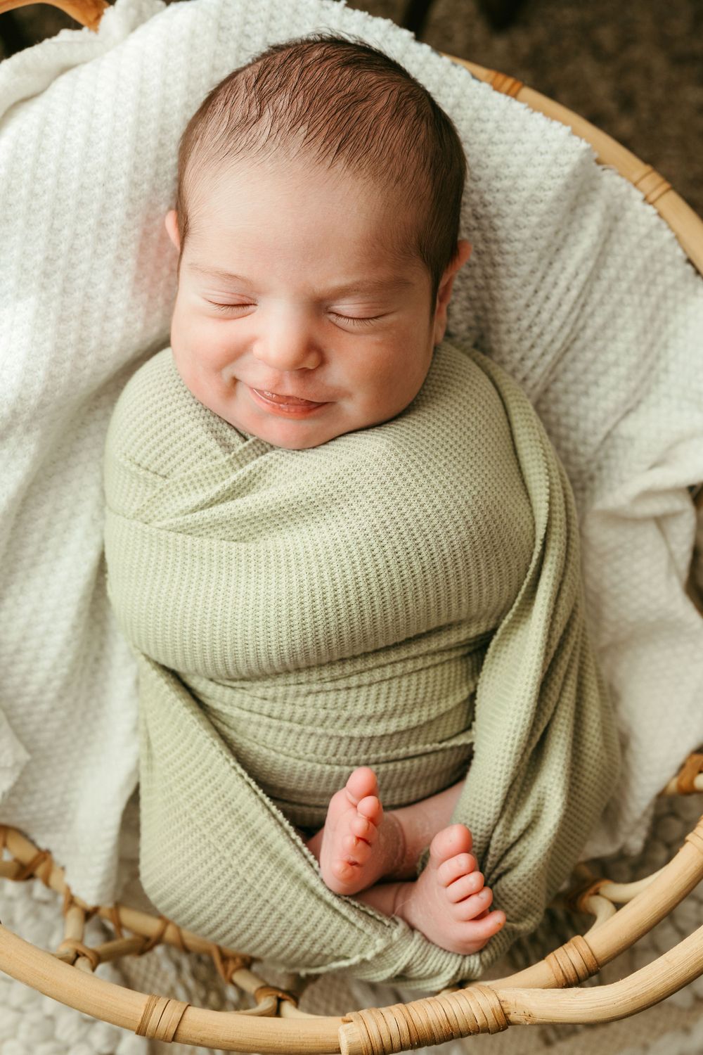 A sleeping newborn wrapped in a sage green knit blanket lies peacefully in a round wicker basket.