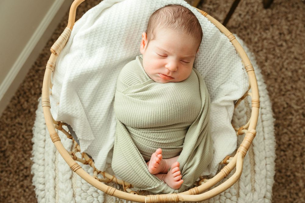 A sleeping newborn wrapped in a sage green swaddle rests peacefully in a round woven basket lined with white fabric.