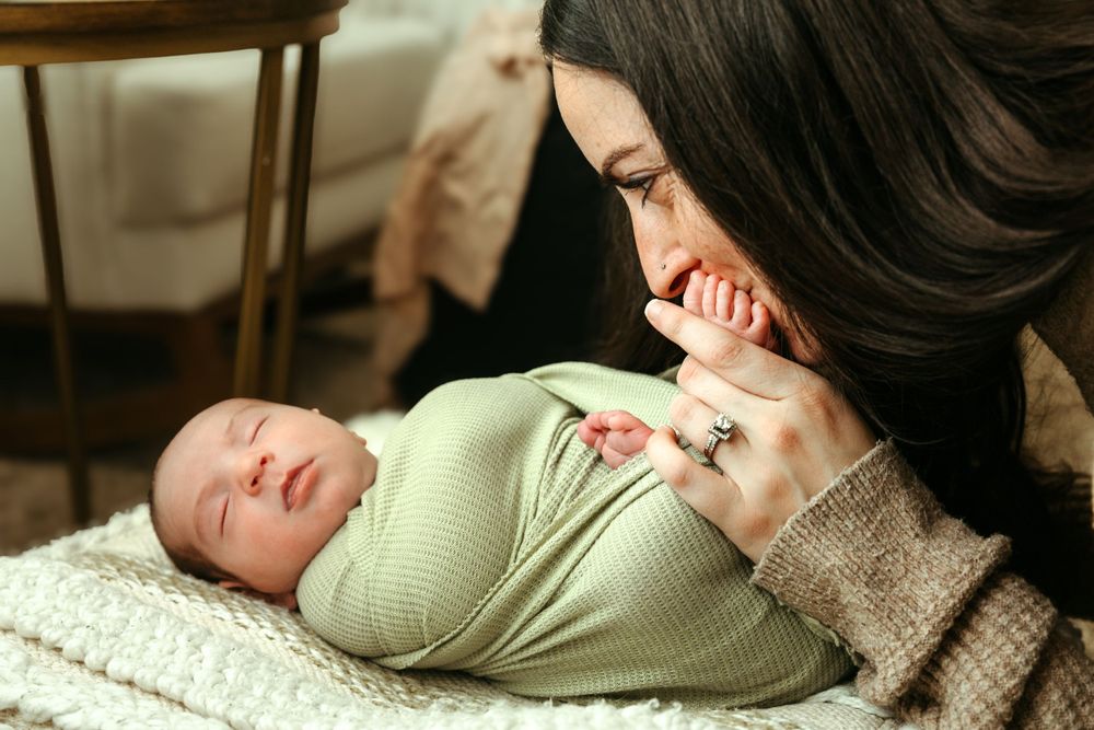 A swaddled newborn baby in mint green sleeps peacefully on a knitted blanket while being watched over with care.