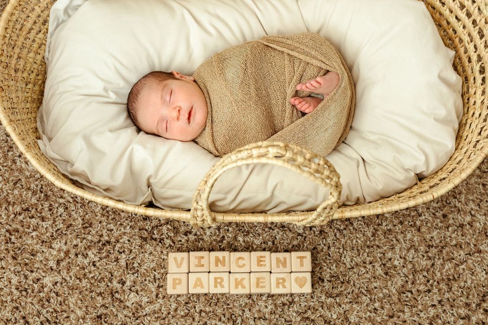 A sleeping infant swaddled in tan fabric lies in a cream-colored basket on brown carpet with wooden letter blocks below.