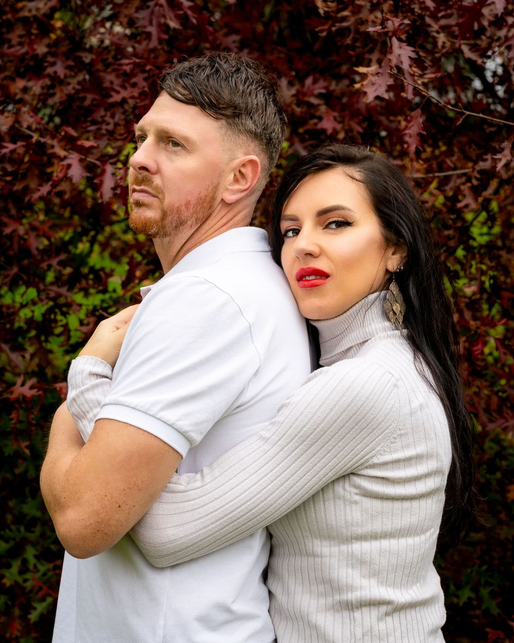 Couple in contrasting white and gray outfits posing against red foliage background.