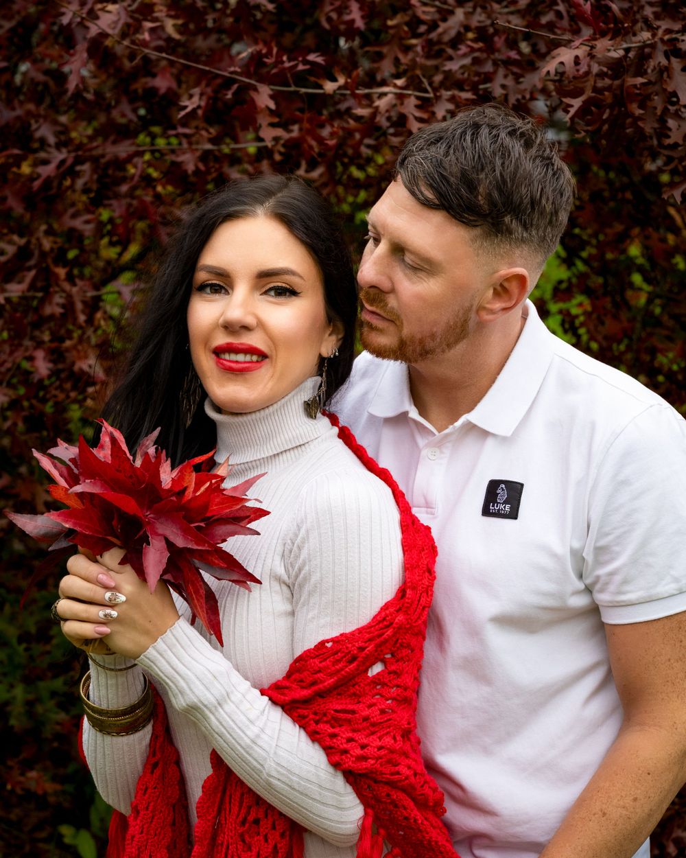 Couple in white outfits posing with red autumn leaf against burgundy foliage.