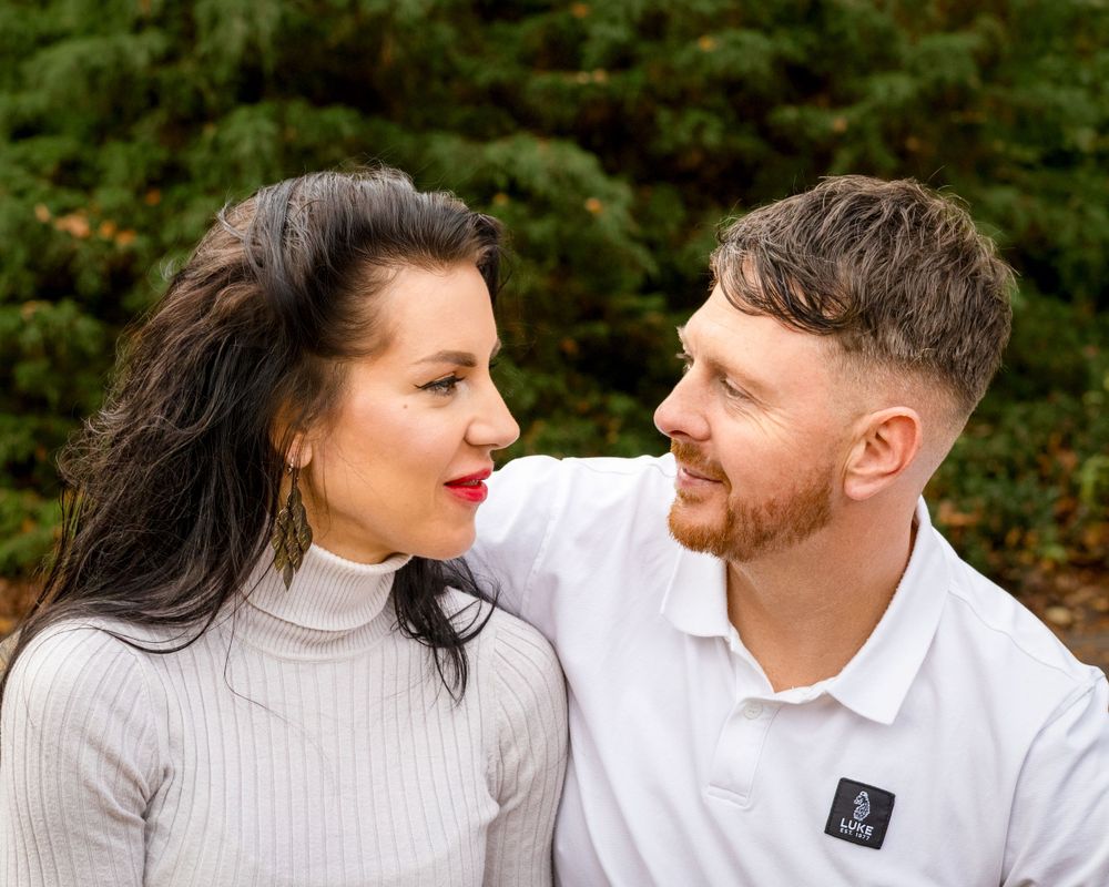 Couple sharing tender moment outdoors in white outfits against green foliage.