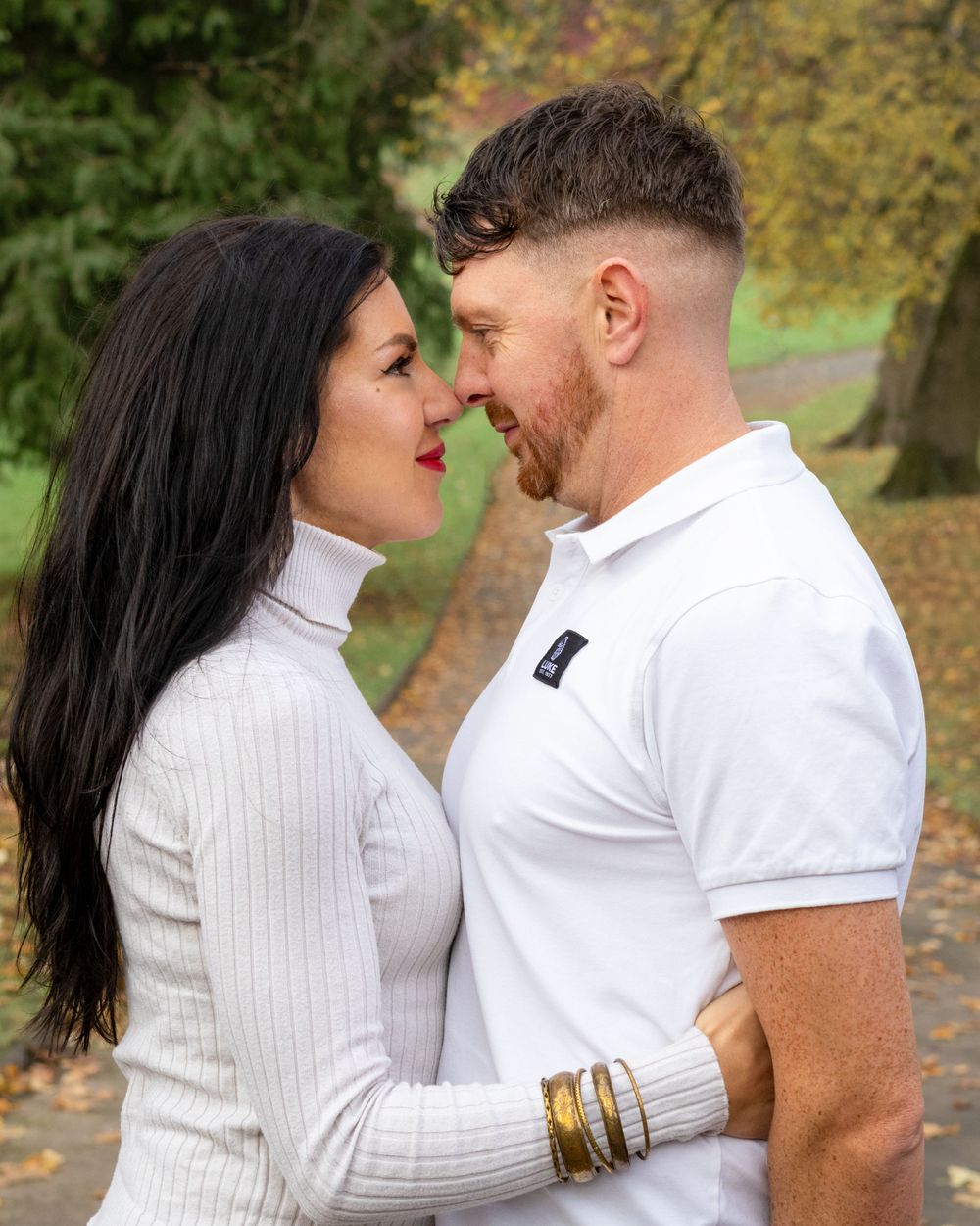 Couple wearing white tops embrace lovingly against fall foliage backdrop.