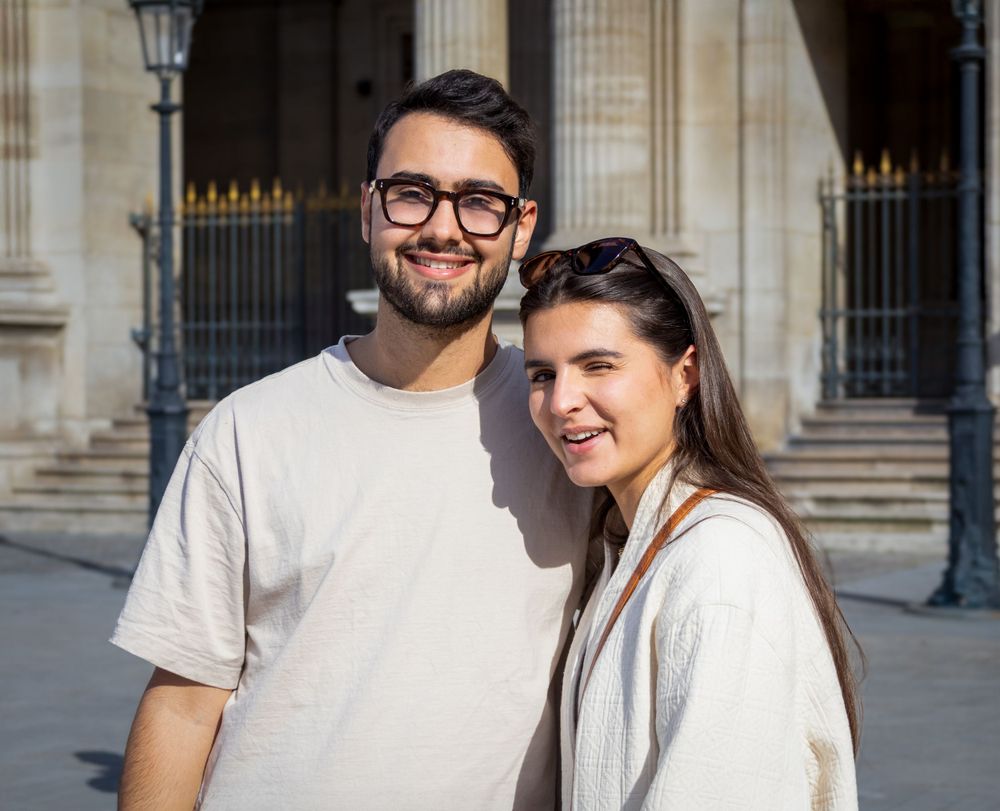 Two smiling people in light colored casual clothing stand together in front of a classical building facade.