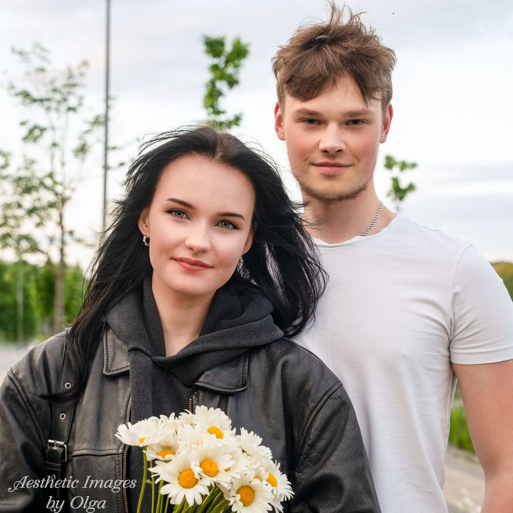 A couple standing outdoors on a cloudy day, with the woman holding white and yellow daisies.