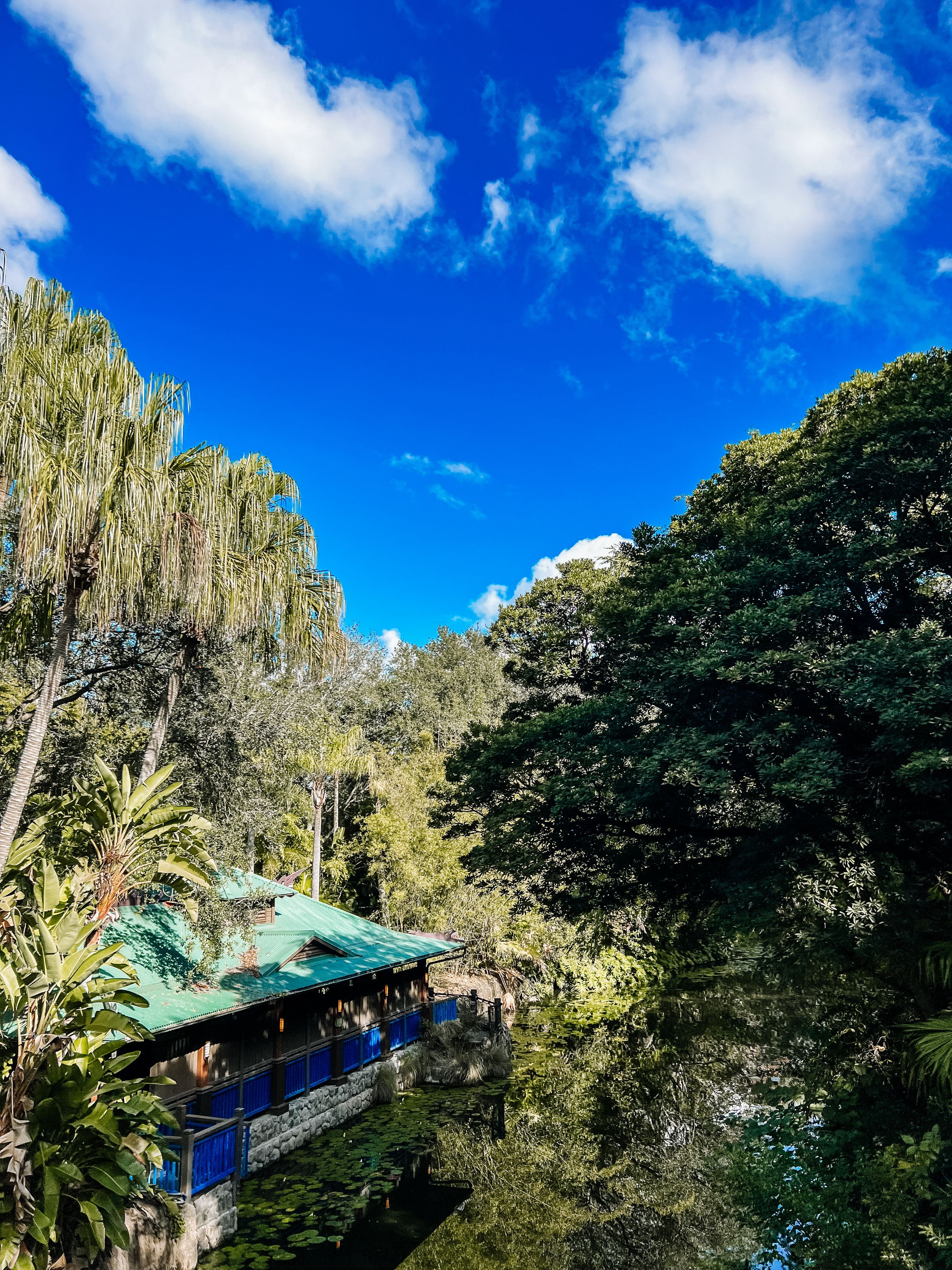 A blue and green pagoda-style structure nestled among lush trees and tropical vegetation under a bright blue sky with clouds.