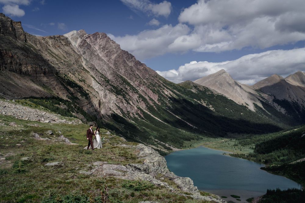 Two hikers stand on a mountain ridge overlooking a turquoise alpine lake surrounded by dramatic peaks in the wilderness.