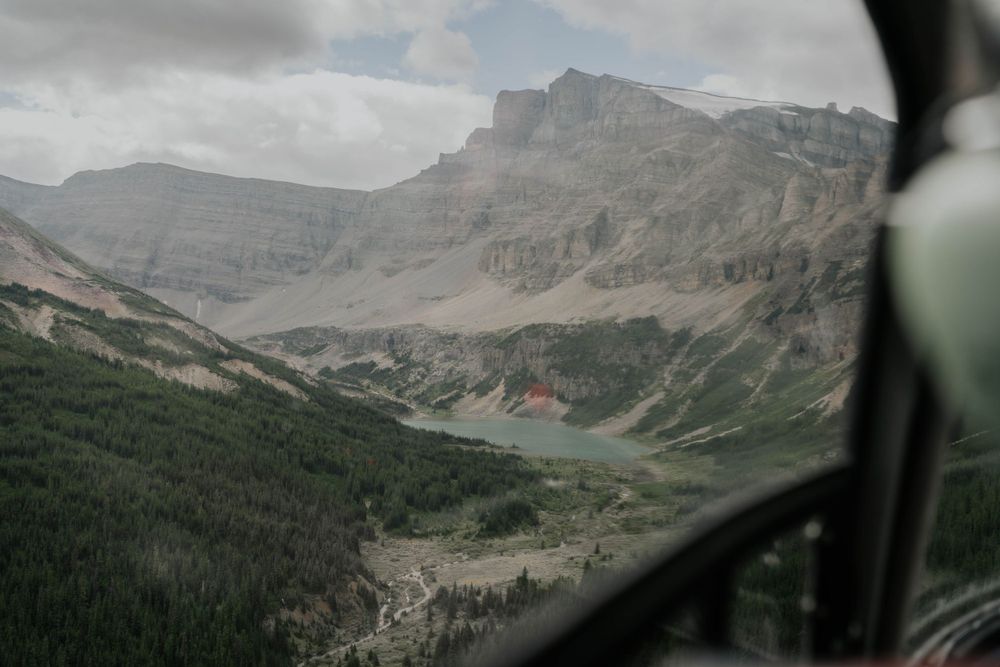 Aerial view through helicopter window of rugged mountain terrain with forest and rocky peaks in the distance.