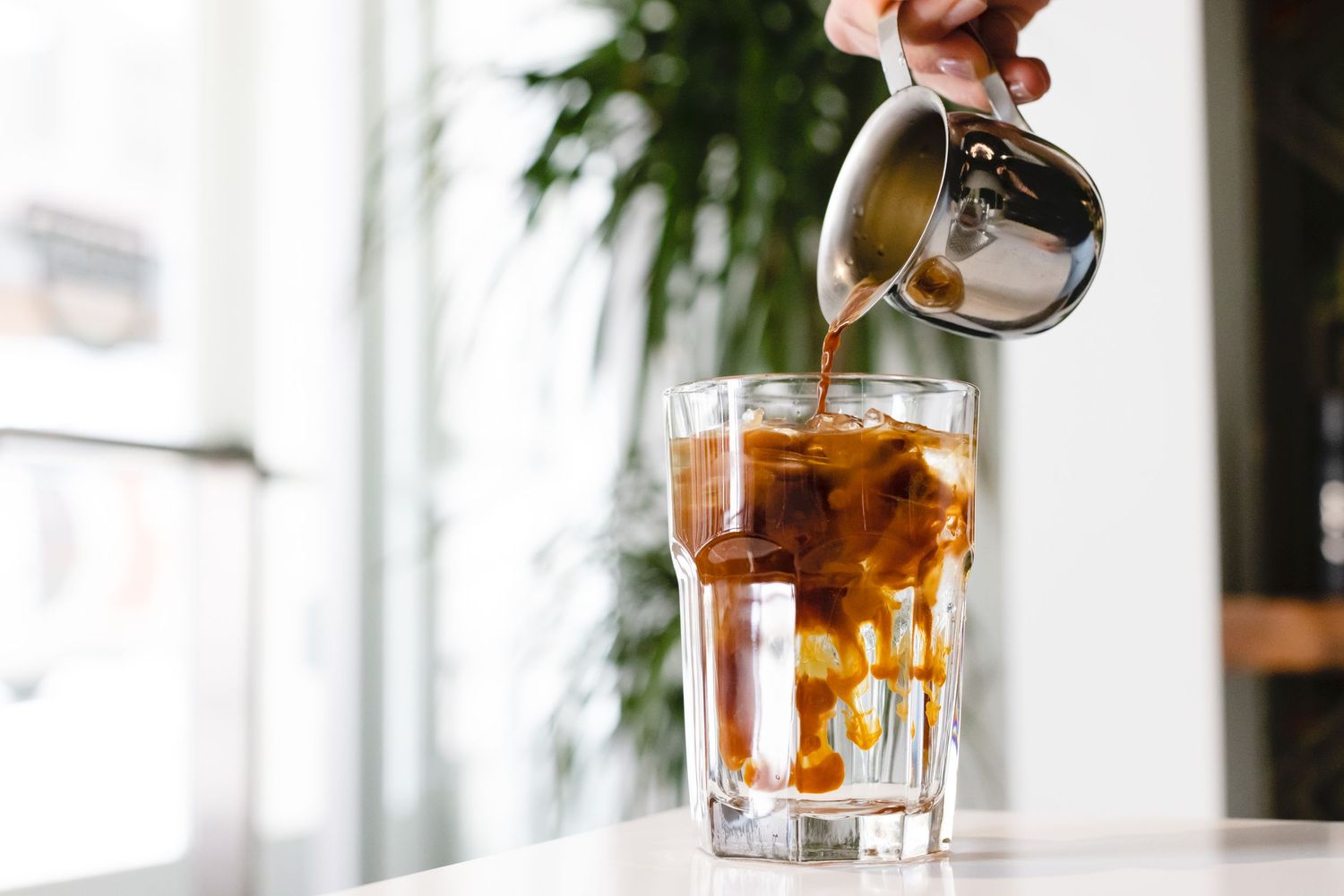 Creamy milk splashes into a glass of iced coffee creating a swirling caramel effect near a bright window.