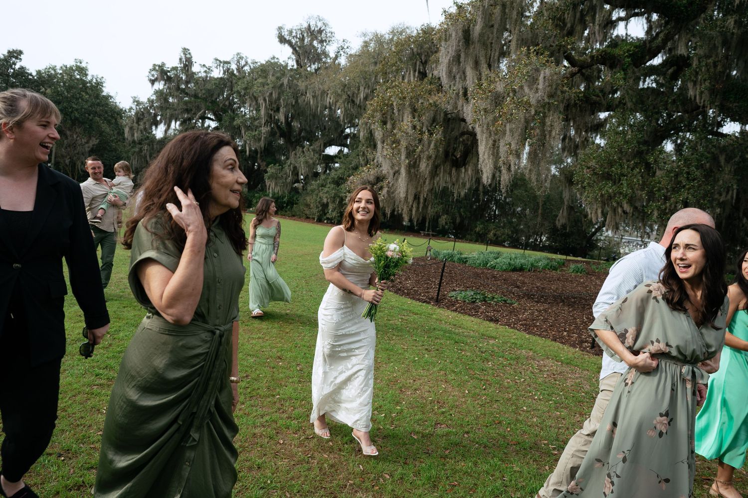 Group of people running and laughing on a green lawn with Spanish moss-draped trees in the background.