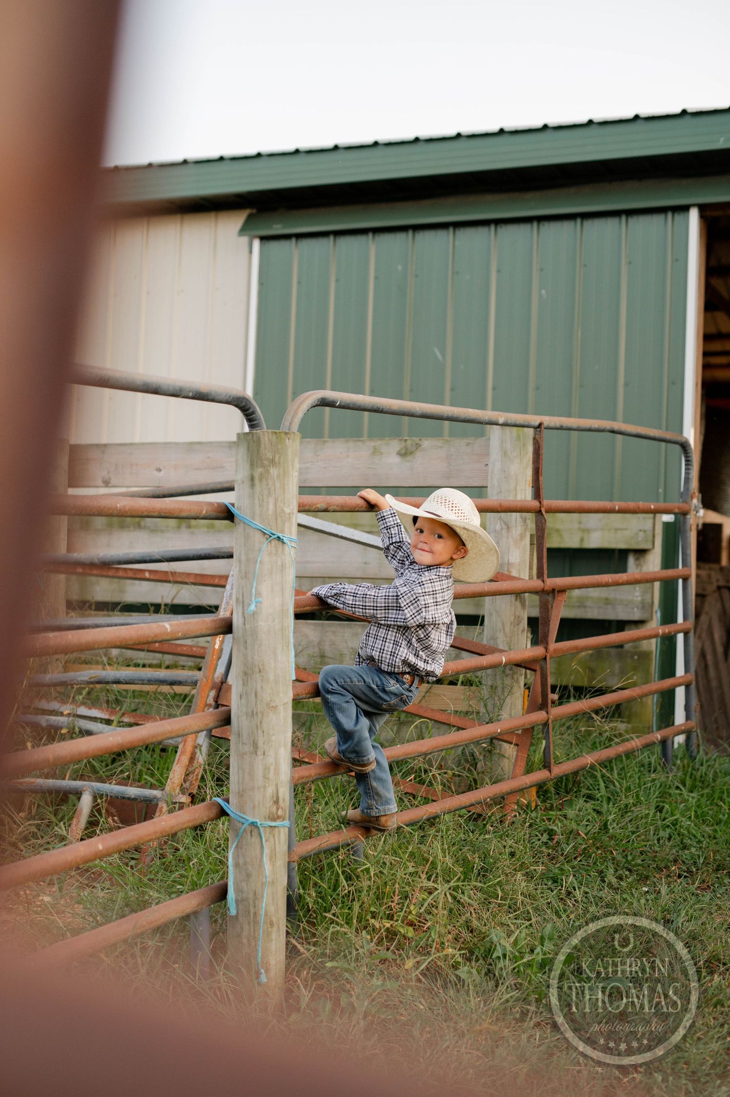 Child in plaid shirt climbing rustic metal fence near barn building.