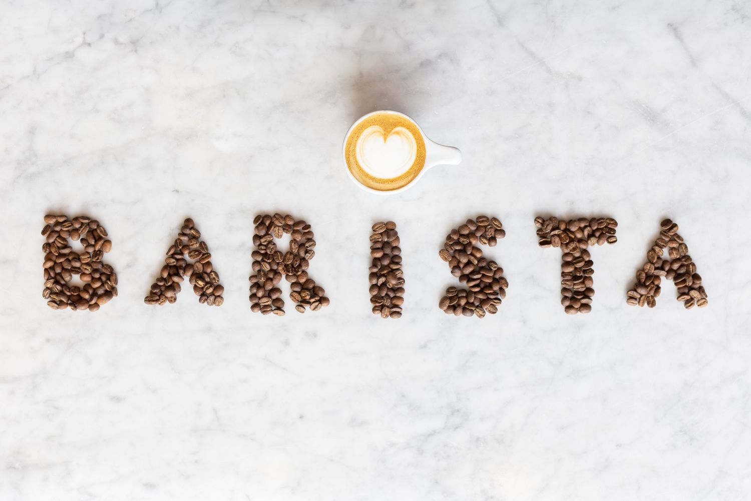 The word 'BARISTA' spelled out in coffee beans on a white surface with a small coffee cup icon above.