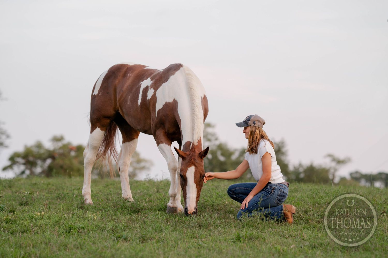 A peaceful scene of someone kneeling beside a brown and white spotted horse grazing in a grassy field.