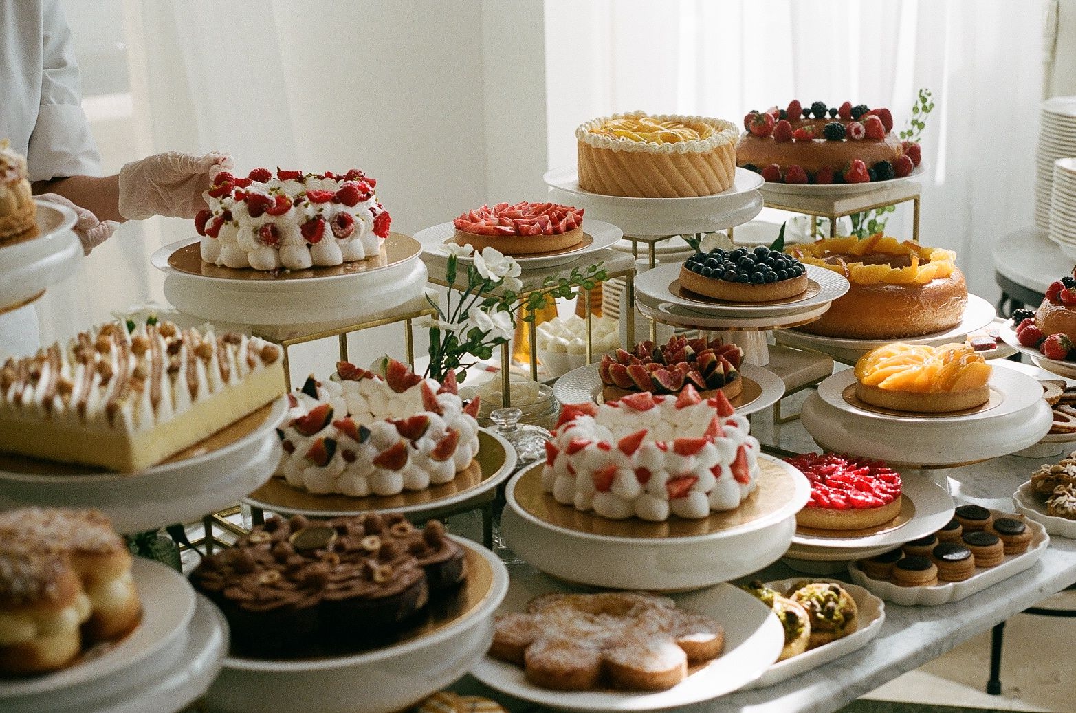 A dessert table filled with various cakes, pastries and fruit-topped treats displayed on tiered white stands and platters.