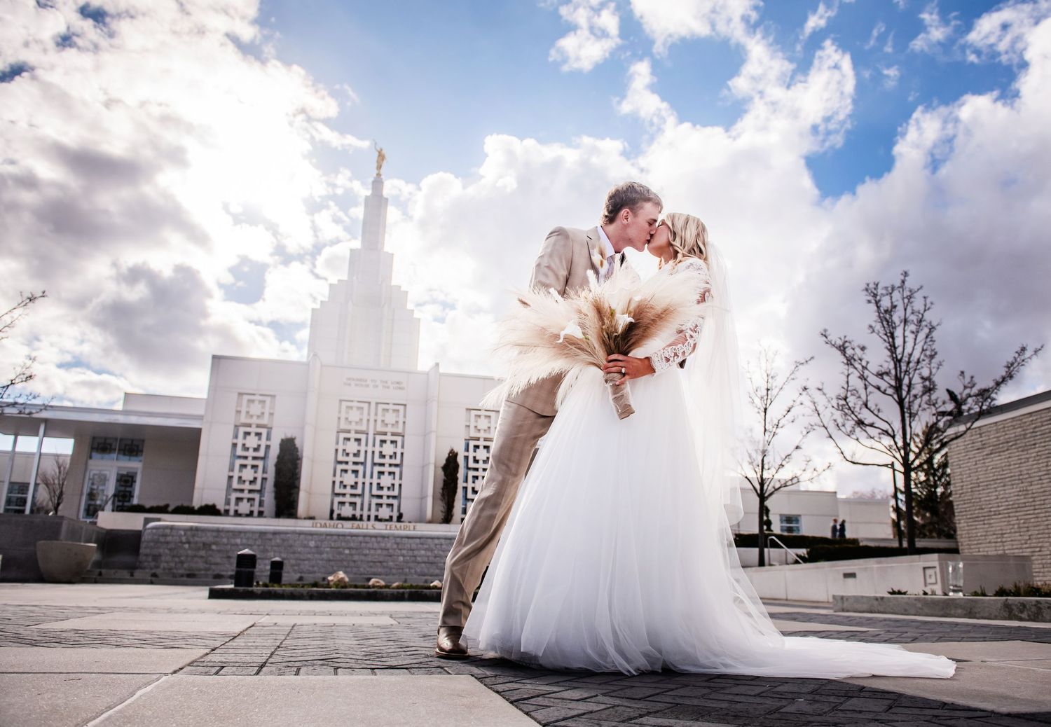 A couple in wedding attire share an intimate moment outside a majestic white church under dramatic cloudy skies.