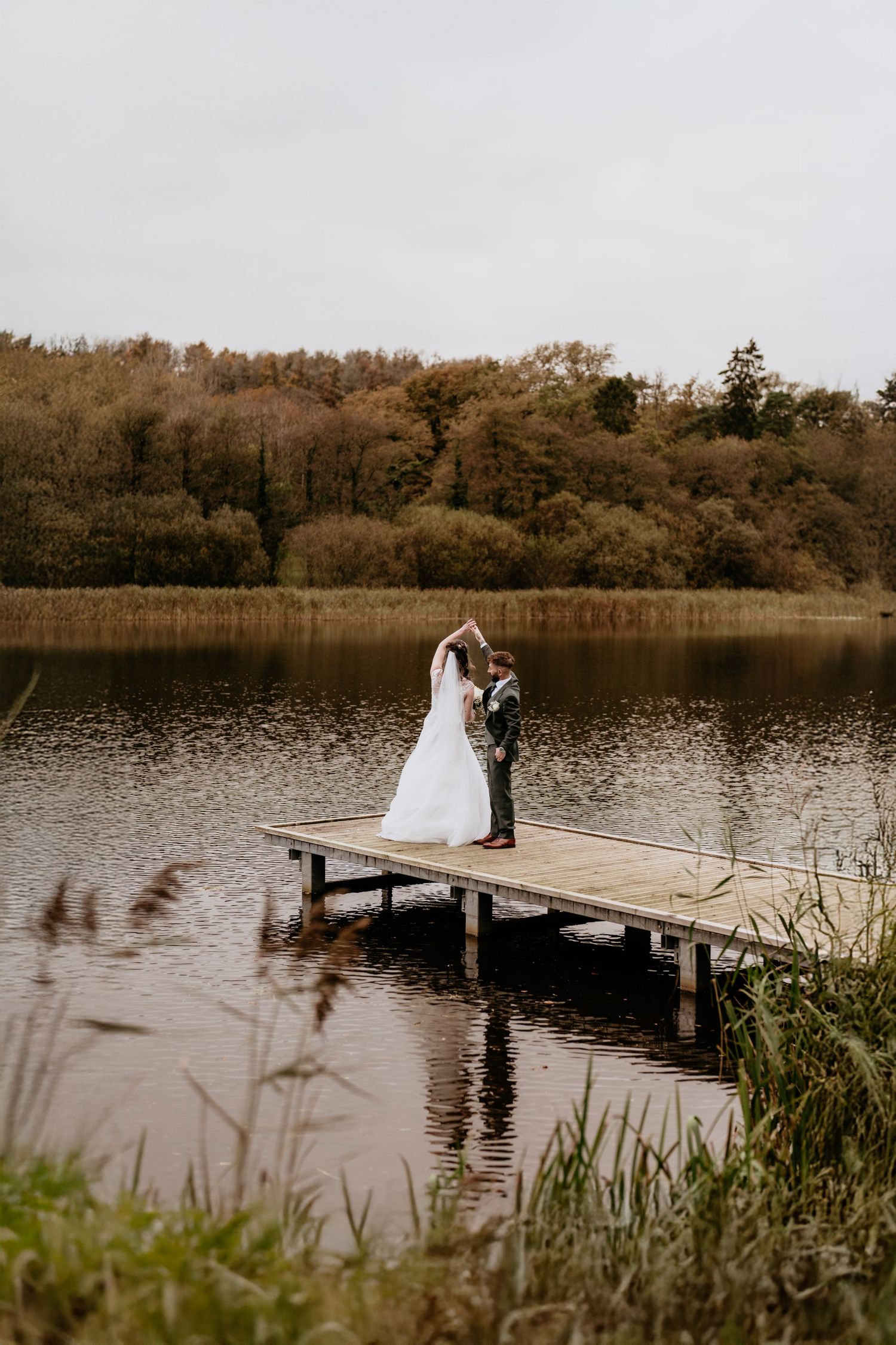 A couple stands together on a wooden dock overlooking a serene lake surrounded by autumn trees on an overcast day.