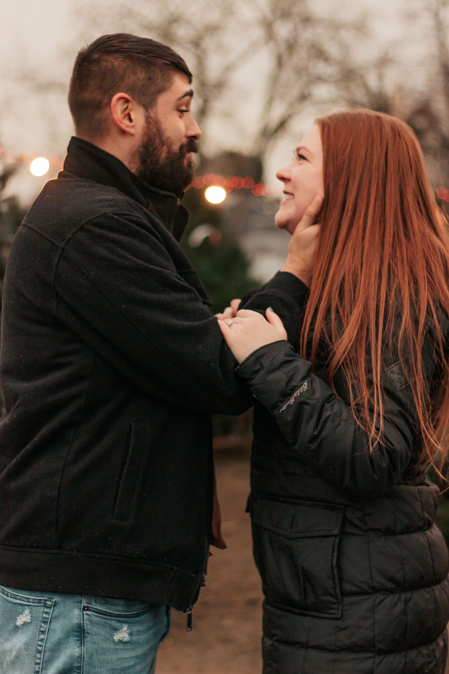 A bearded man in a black jacket holds the chin of a smiling redhead woman outdoors at dusk | Westminster Colorado Proposal Photographer