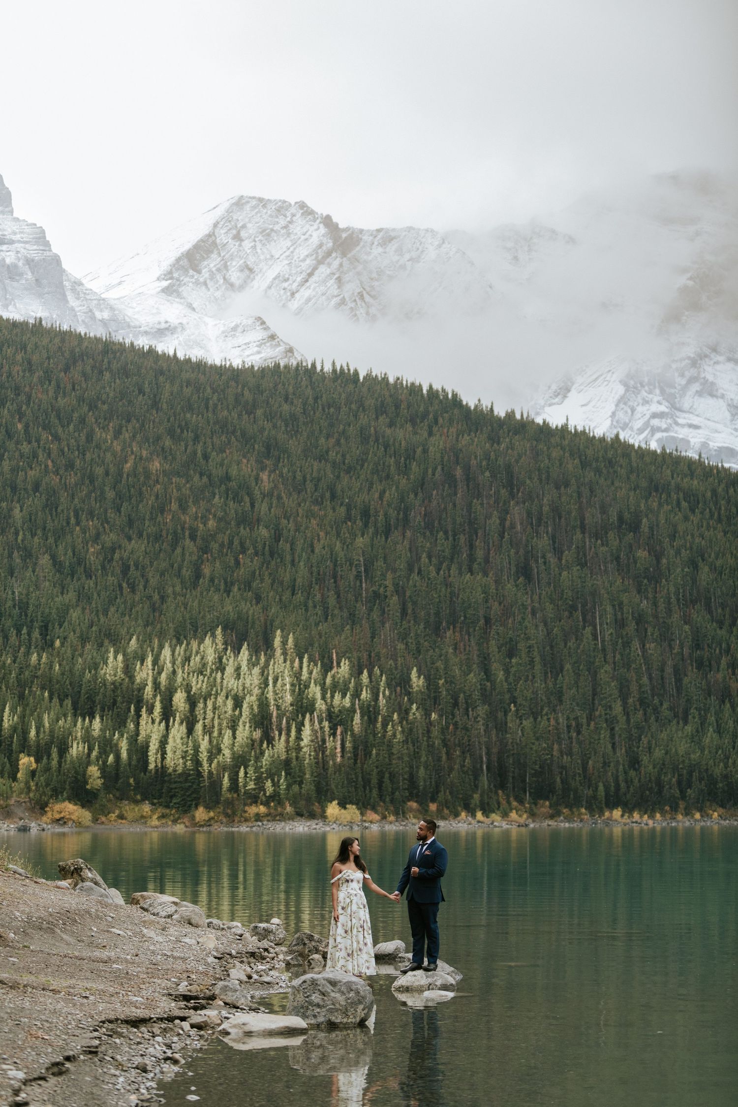 A couple stands on rocks at the edge of a serene mountain lake with misty peaks in the background.